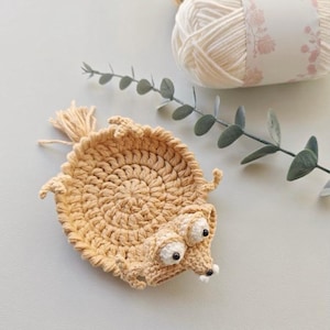 Peut inclure: Un dessous de verre en crochet beige en forme de petit animal avec un corps rond, quatre membres courts, une queue et une t&ecirc;te dot&eacute;e de deux grands yeux blancs &agrave; pupilles noires et d'un petit museau. L'objet est fait de fil &eacute;pais en cercle.