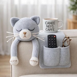 Puede incluir: Un organizador de brazo de sofá con forma de gato de ganchillo gris claro con una taza que dice "CANEE NEKS CAPFE", un mando a distancia y gafas. El gato tiene los ojos cerrados, bigotes blancos y patas blancas. El organizador tiene dos bolsillos.