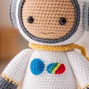 Könnte beinhalten: Handgefertigte Astronautenpuppe aus Häkelarbeit. Hauptsächlich weiß mit grauen und gelben Akzenten. Das Gesicht ist beige mit schwarzen Augen und einem Lächeln. Auf der Brust befinden sich ein blauer Kreis und ein Regenbogen-Patch.