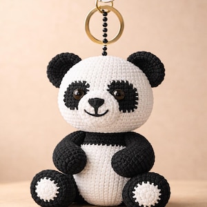 Op de afbeelding: Een handgemaakte panda beer sleutelhanger. De panda is gehaakt met wit en zwart garen, met zwarte oren, ogen en ledematen. Hij heeft een lachend gezicht en is bevestigd aan een zilveren en gouden sleutelhanger.