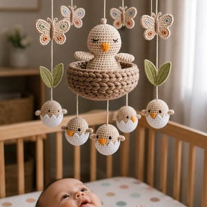 May include: A handcrafted baby mobile with a crocheted chick in a nest, butterflies, and hatching eggs. The mobile hangs above a baby in a cot. The colours are soft and neutral, with beige, white, and light green accents.