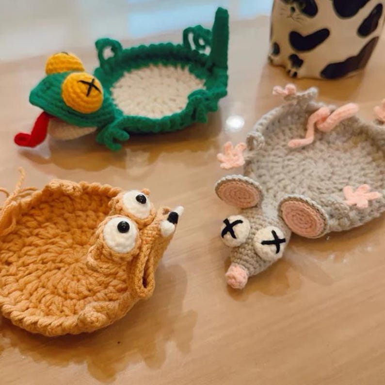 May include: A collection of whimsical, handmade crochet coasters. The coasters feature unique animal designs, including a frog, a rat, and a creature with large eyes. Each coaster has distinct colors and details, adding a playful touch to any home decor.