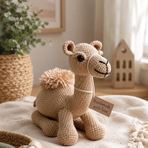 Puede incluir: Un juguete de camello tejido a ganchillo en posición sentada, hecho de hilo de color beige claro. El camello tiene ojos pequeños y oscuros, una joroba esponjosa y una etiqueta que dice "Handmade with love". Está sobre una manta crema.