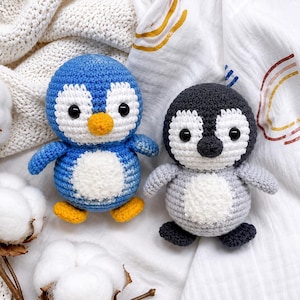 May include: Two crocheted penguin plush toys. One penguin is blue and white with a yellow beak and feet. The other penguin is dark gray, light gray, and white with a black beak and feet. Both have black plastic eyes and round white bellies.