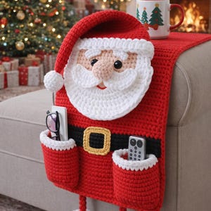 May include: A red and white crocheted Santa Claus couch organizer with pockets for glasses and a remote. The Santa design includes a white beard, hat, and trim, with a black belt and gold buckle. The organizer is placed on a couch arm.
