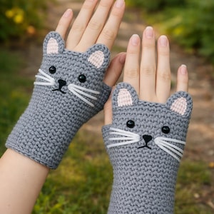Puede incluir: Un par de guantes sin dedos de ganchillo grises con forma de gatos. Los guantes tienen ojos negros, bigotes blancos y orejas internas rosas. Los guantes están diseñados para cubrir la mano hasta los nudillos.