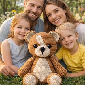 Può includere: Un orsacchiotto all'uncinetto con filato marrone e crema, seduto sull'erba verde. L'orso ha una striscia bianca sulla testa e un naso nero. Una famiglia di quattro persone sorride sullo sfondo.