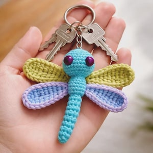 May include: A crocheted dragonfly keyring featuring a light blue body and head with two dark purple eyes. The dragonfly has two pairs of wings, with the upper pair in light green and the lower pair in light purple, attached to a metal ring.