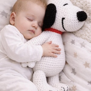 May include: A sleeping baby cuddles a handmade white crochet Snoopy plush toy. The Snoopy has black ears and nose, a red collar, and is lying on a star-patterned blanket. The baby is wearing a white onesie.