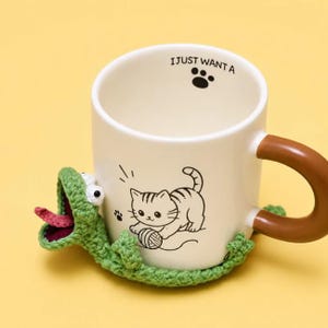 Peut inclure: Une tasse en c&eacute;ramique blanche avec une anse marron, orn&eacute;e d'un dessin au trait noir repr&eacute;sentant un chaton jouant avec une pelote de laine. Le bord int&eacute;rieur porte le texte I JUST WANT A et une empreinte de patte. Un l&eacute;zard crochet&eacute; vert entoure la base.