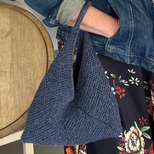 May include: A triangular, navy blue knitted tote bag with a handle. The bag is held by a person wearing a denim jacket and a floral print dress. The background includes a wooden barrel.
