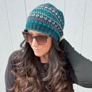 May include: A teal blue crochet beanie with a white, pink, and green geometric pattern. The beanie is slouchy and has a pompom on top.