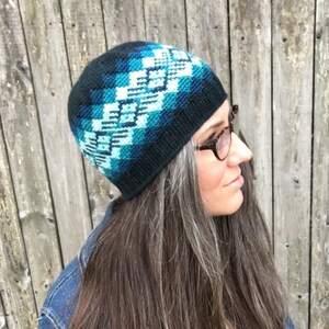 May include: A black and blue knitted beanie with a geometric pattern. The beanie is being worn by a person with long, grey hair.