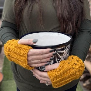 May include: A woman wearing sunglasses and mustard yellow fingerless gloves holds a decorative ceramic bowl with a black and white design. The bowl is white inside. The woman is wearing a green long-sleeve shirt.