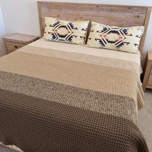 Pode incluir: Uma cama com cabeceira de madeira, duas mesinhas de cabeceira a condizer e um cobertor em camadas. O cobertor transita de bege claro para bege médio e castanho escuro. Duas almofadas decorativas com padrões geométricos estão na cama.