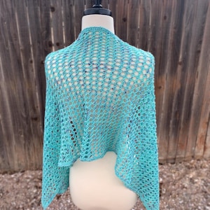 Puede incluir: Un chal de ganchillo turquesa exhibido en un maniquí. El diseño de tejido abierto del chal es visible, con una apariencia ligeramente texturizada. El chal está drapeado sobre los hombros del maniquí, mostrando su forma y caída. El fondo es una valla de madera.