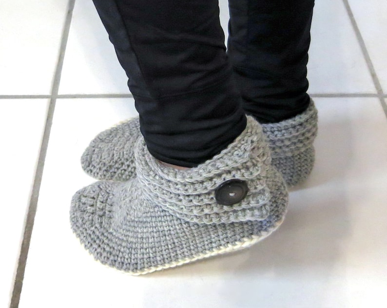 Pu&ograve; includere: Stivali da camera in crochet grigi con un dettaglio a bottone. Gli stivali hanno un risvolto piegato e un bordo bianco.