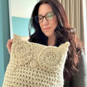 May include: A crocheted owl pillow in a cream color. The pillow is being held by a person wearing glasses.