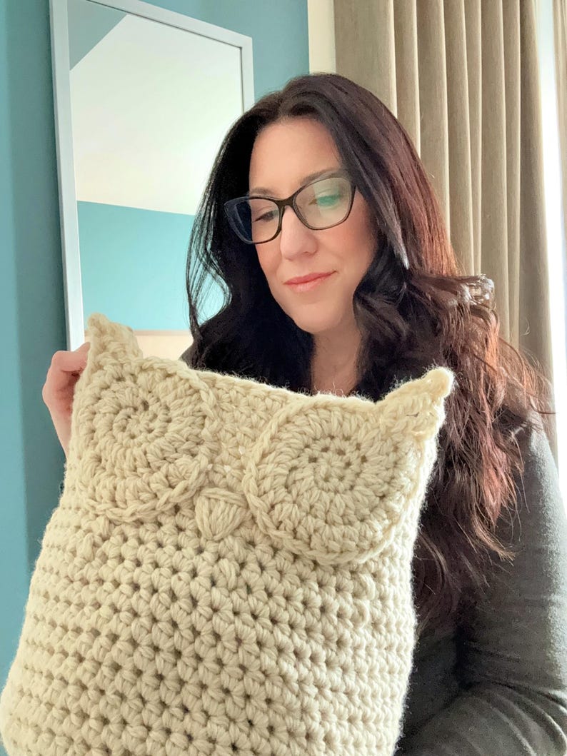 May include: A crocheted owl pillow in a cream color. The pillow is being held by a person wearing glasses.