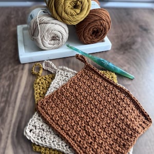 May include: A collection of handmade crochet items in warm, earthy tones. The image features several skeins of yarn in mustard yellow, brown, and beige, along with three crocheted potholders and a crochet hook. The potholders are stacked on a wooden surface.