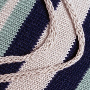 May include: Close-up of a knitted fabric with stripes of beige, blue, and green. The fabric is woven in a diagonal pattern, creating a textured look.
