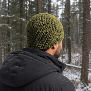May include: A green knitted beanie hat. The hat is a solid olive green color and has a textured, woven appearance. The person is wearing a black jacket. The background is a snowy forest.