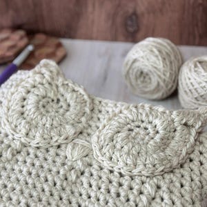 May include: A beige crocheted bag with two large circular designs on the front. The bag is made with a chunky yarn and has a simple, elegant design.