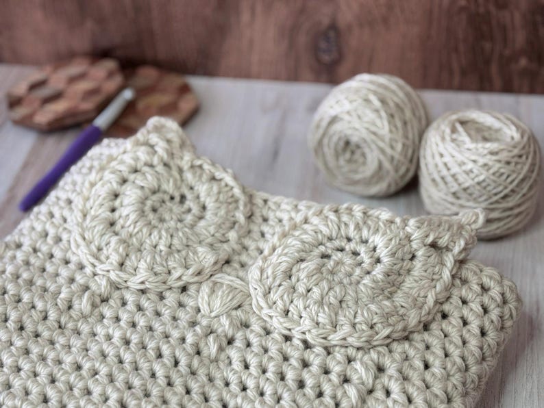 May include: A beige crocheted bag with two large circular designs on the front. The bag is made with a chunky yarn and has a simple, elegant design.