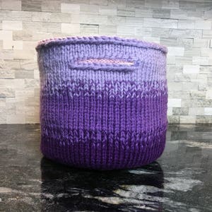 May include: A purple and lavender ombre knitted storage basket with a handle.