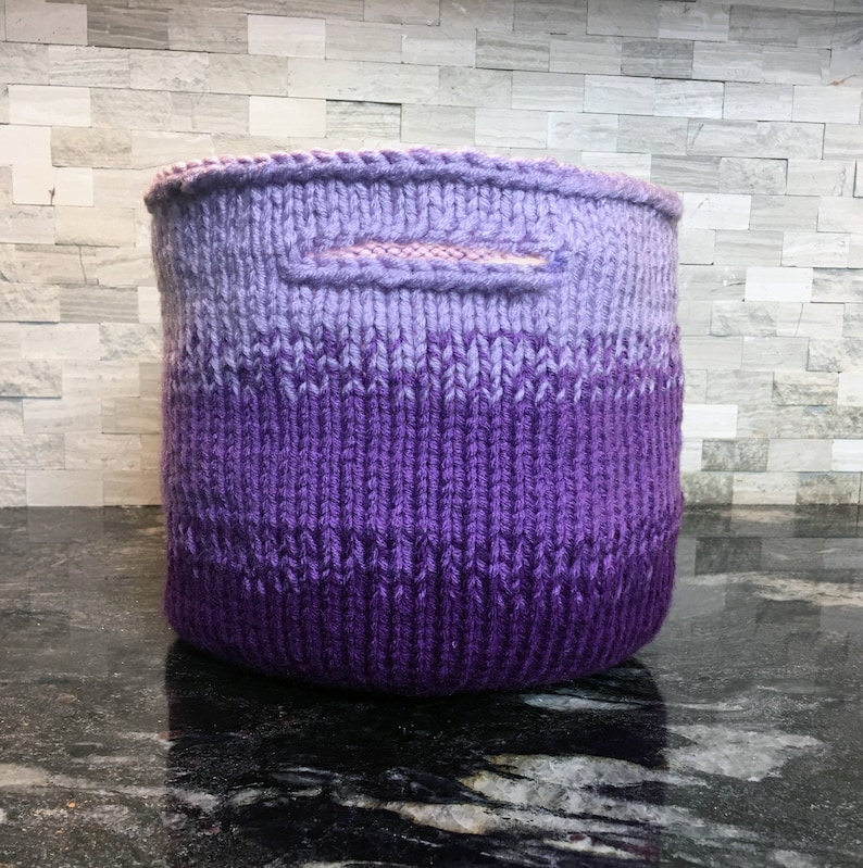 May include: A purple and lavender ombre knitted storage basket with a handle.