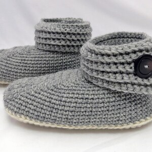 Pu&ograve; includere: Ciabatte in crochet grigio con polsino ripiegabile e un bottone nero. Le ciabatte hanno un bordo bianco attorno alla suola.