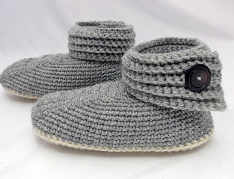Pu&ograve; includere: Ciabatte in crochet grigio con polsino ripiegabile e un bottone nero. Le ciabatte hanno un bordo bianco attorno alla suola.