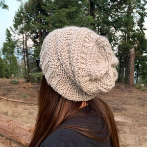 May include: A beige knitted beanie with a slouchy fit. The beanie is made of a soft, textured yarn.