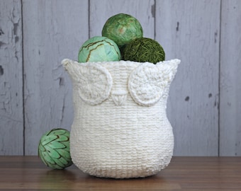 May include: A white crocheted owl-shaped basket filled with green decorative balls. The basket is sitting on a wooden surface.