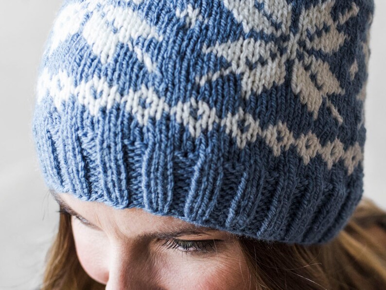 Pu&ograve; includere: Un cappello di lana blu e bianco con un motivo geometrico. Il cappello &egrave; indossato da una persona con i capelli castani.