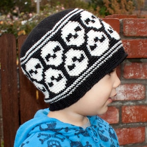 May include: A child wearing a black and white skull patterned knit hat. The hat has a white brim and is crocheted. The child is wearing a blue shirt with a skull pattern.