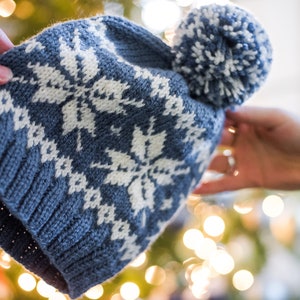 Pu&ograve; includere: Un cappello di lana blu con un motivo a fiocco di neve bianco e un pon pon bianco in cima.