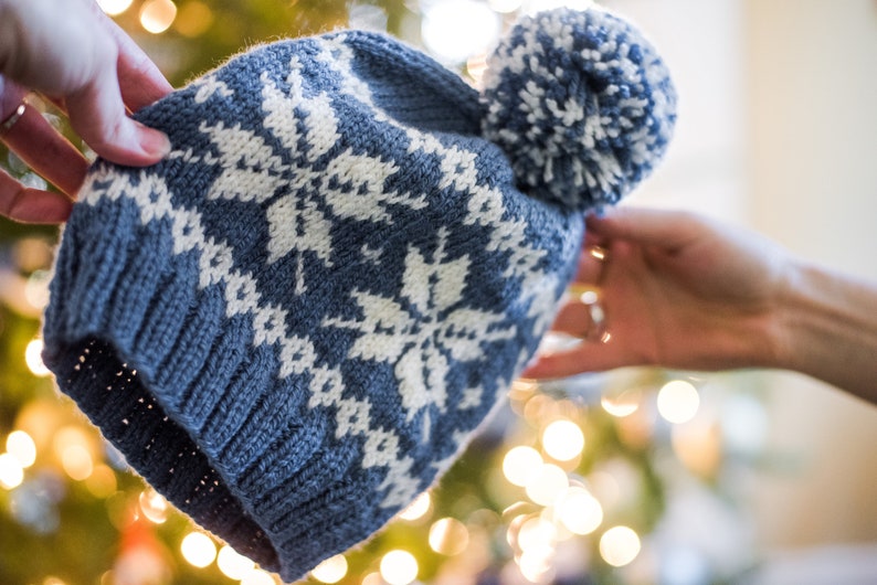 Pu&ograve; includere: Un cappello di lana blu con un motivo a fiocco di neve bianco e un pon pon bianco in cima.