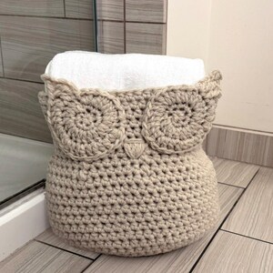 May include: A crocheted owl-shaped basket in beige with a white towel inside. The basket is sitting on a tiled floor.