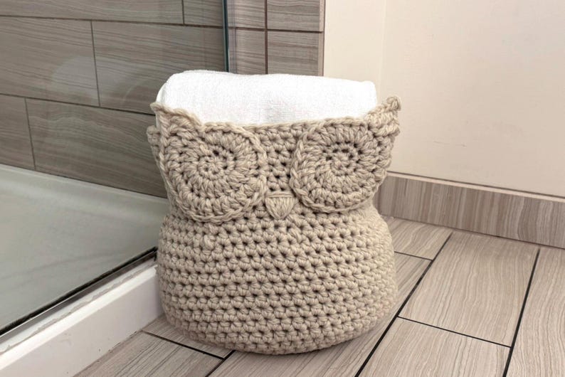 May include: A crocheted owl-shaped basket in beige with a white towel inside. The basket is sitting on a tiled floor.