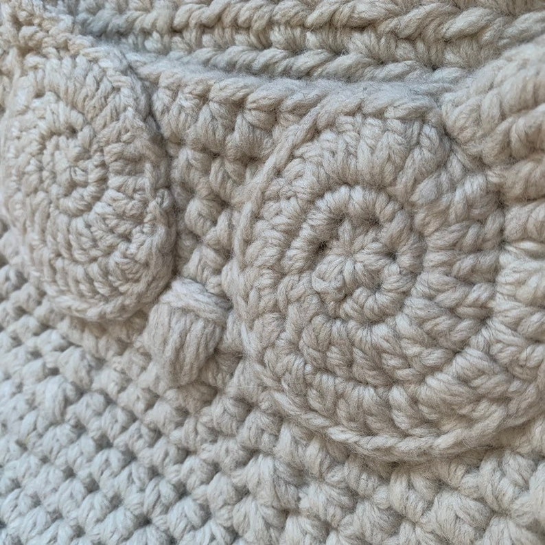 May include: Close-up of a beige crocheted fabric with a textured pattern of circles. The fabric is made with thick yarn and has a soft, fuzzy texture.