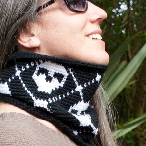 May include: A black and white crocheted neck warmer with a geometric pattern. The neck warmer is being worn by a person with grey hair.