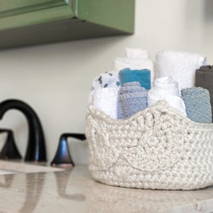 May include: A white crocheted basket filled with a variety of towels in shades of blue, white, and grey. The basket is sitting on a white countertop.