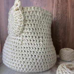 May include: A beige crocheted basket with a decorative crocheted detail on the side. Two balls of beige yarn are in the foreground.
