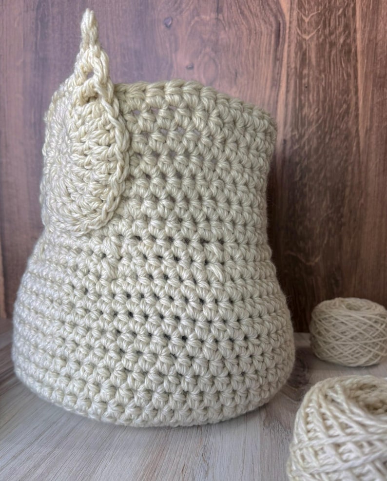 May include: A beige crocheted basket with a decorative crocheted detail on the side. Two balls of beige yarn are in the foreground.