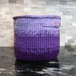 May include: A purple and lavender ombre knitted basket with a pink fabric handle.