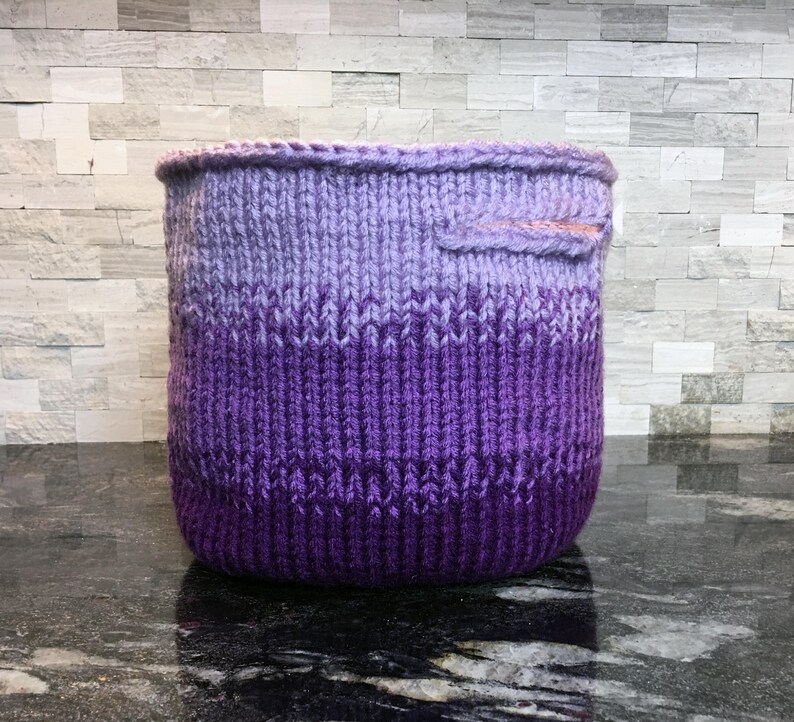 May include: A purple and lavender ombre knitted basket with a pink fabric handle.