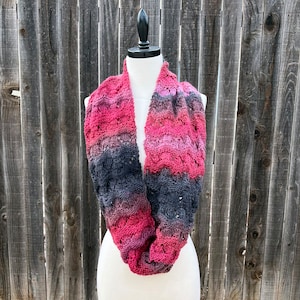 May include: A knitted infinity scarf with a gradient of pink and gray colors. The scarf is displayed on a white mannequin against a wooden fence background.