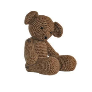May include: A brown crocheted teddy bear sitting with its legs crossed. The bear has black eyes and a brown nose.