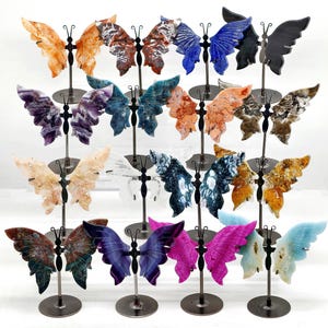 May include: A display of gemstone butterfly sculptures in a range of colours, such as orange, brown, blue, purple, and green. Each butterfly is mounted on a black metal stand, highlighting the detailed carving of the wings.